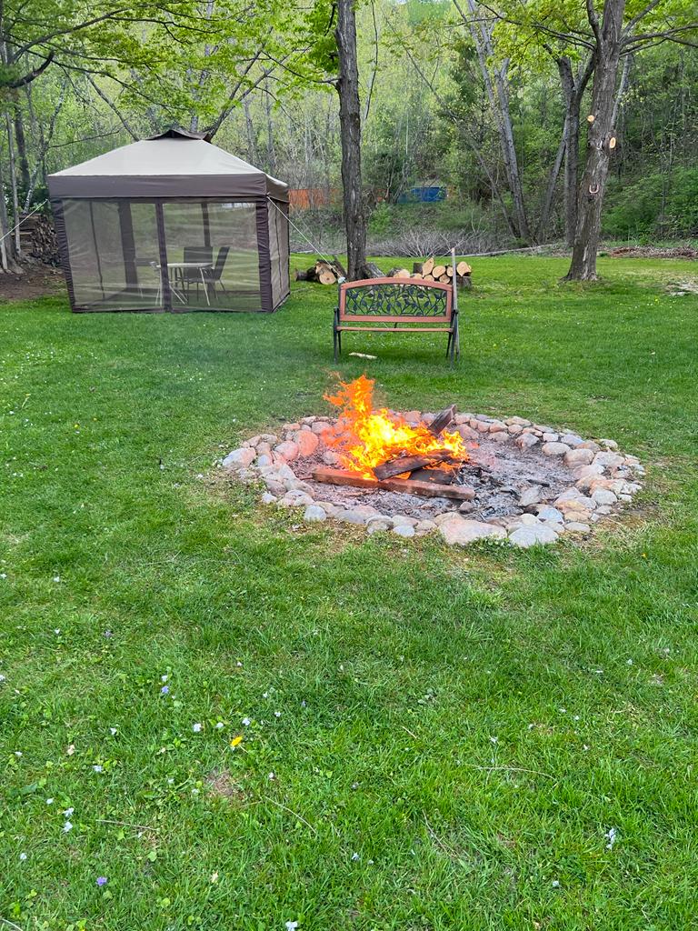 A cozy fire pit surrounded by stone is located on a grassy area, with vibrant flames visible. A wooden bench is positioned nearby, and a gazebo with a screened-in area is seen in the background, providing comfort for outdoor gatherings.
