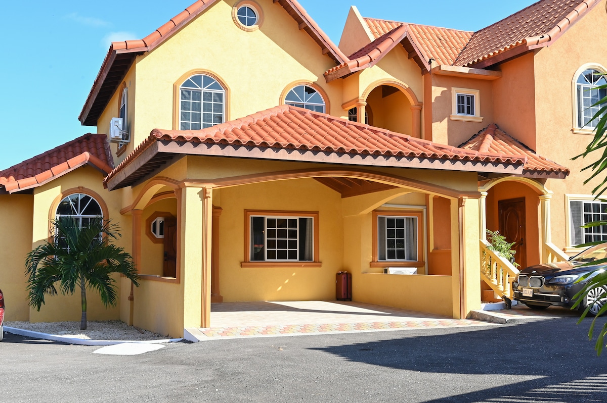 The exterior of the villa is showcased, featuring a vibrant yellow facade with a tiled roof. A covered parking area is visible, along with a well-maintained pathway. Lush greenery is present alongside the entrance, creating a welcoming approach to the property.