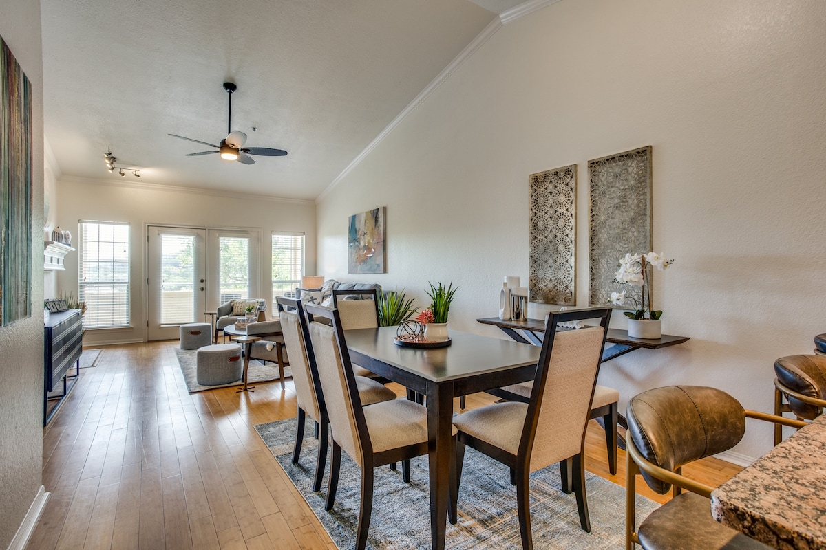 An open dining area features a dark wood dining table surrounded by light-colored chairs, set against a backdrop of wood flooring. Natural light is let in through large windows, enhancing the spacious feel of the room. Decorative wall art adds a touch of elegance.