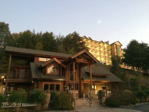 A charming lodge with a log cabin design features large windows and a sloped roof. Surrounding greenery enhances its rustic appeal. In the background, a multi-story building is visible, illuminated by soft lighting as dusk sets in, contrasting with the natural landscape.