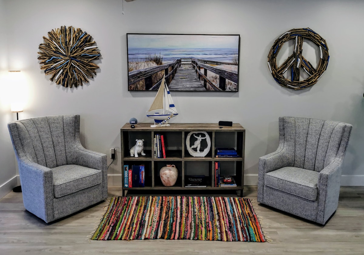 A cozy seating area features two upholstered chairs flanking a low bookshelf filled with books and decorative items. A colorful rug lies beneath, complementing a wall art piece depicting a serene beach scene, with nautical decor adding warmth to the space.