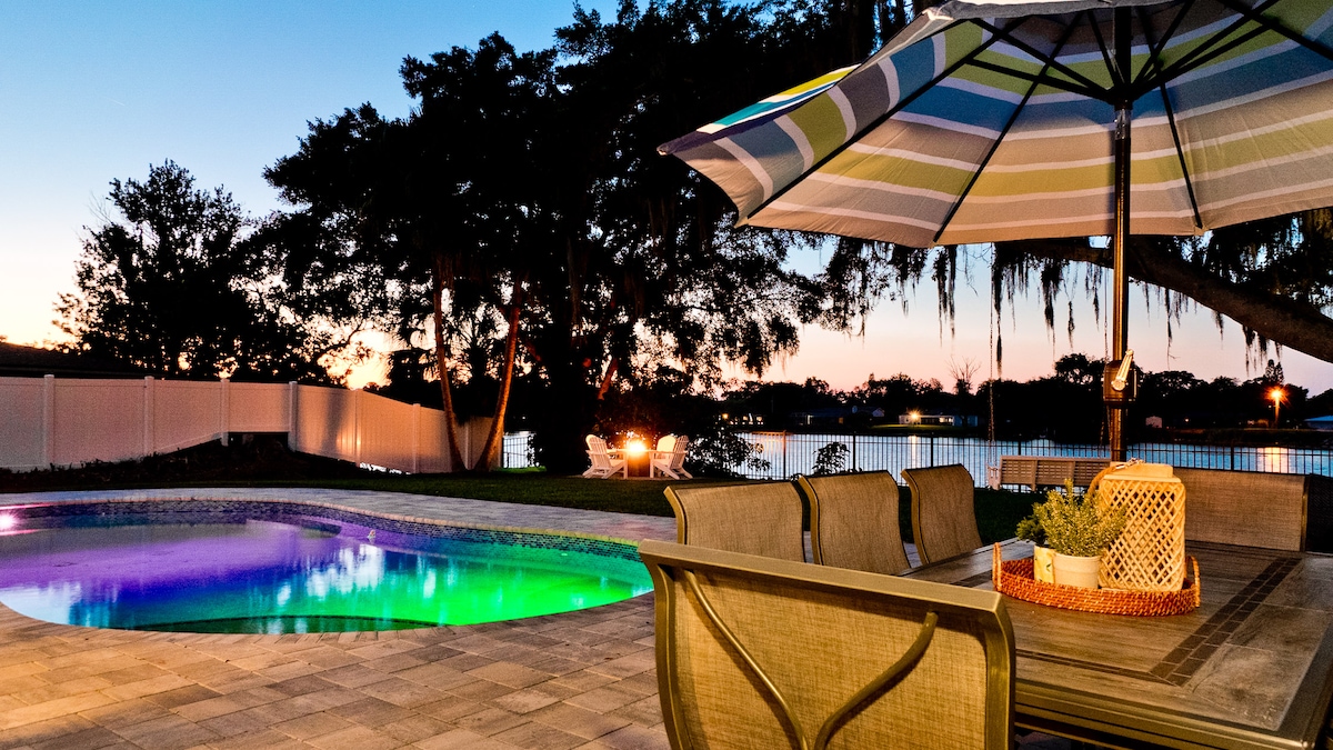 A tranquil backyard is highlighted by a heated saltwater pool, illuminated with soft colored lights. A spacious dining area with a table and chairs sits beneath a large umbrella, providing shade. The serene lake view is framed by trees and a peaceful sunset sky.
