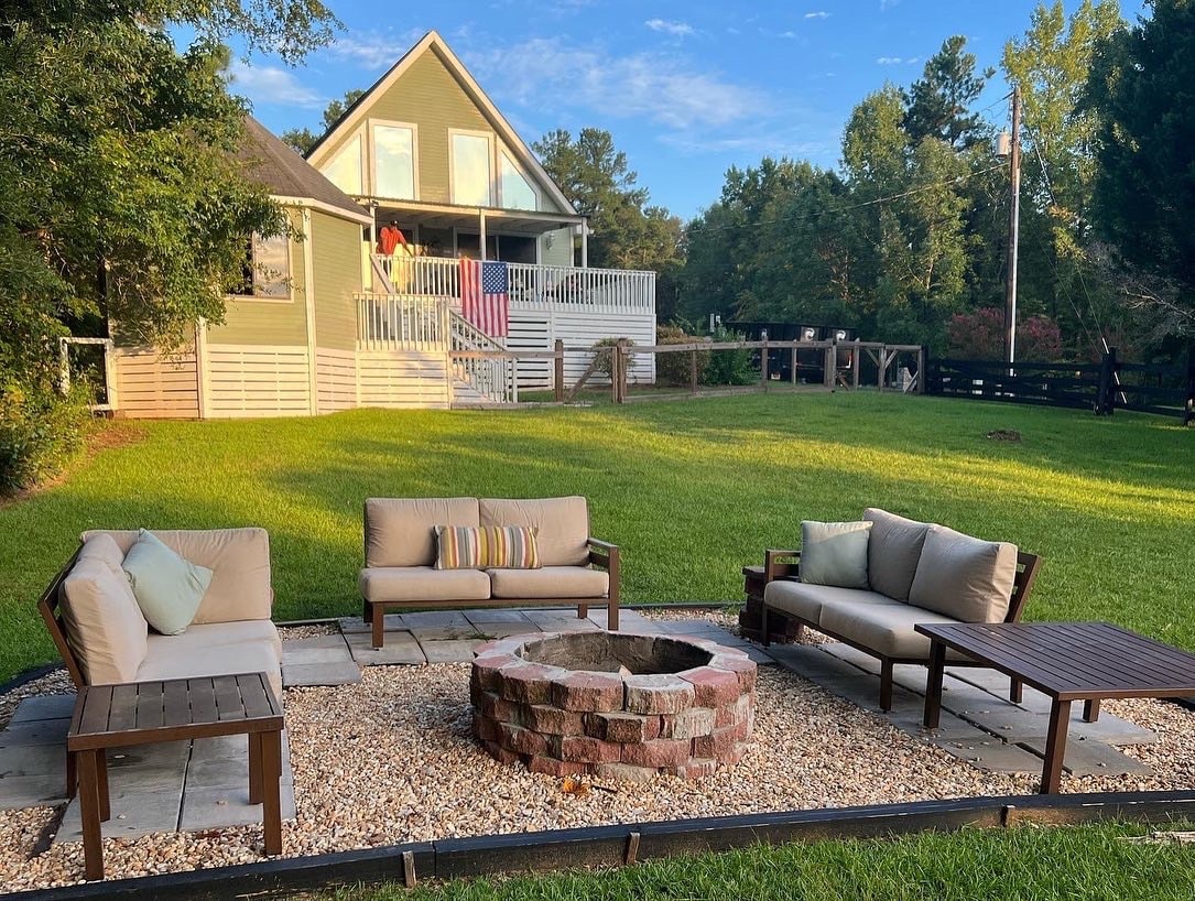 A spacious outdoor area features a circular firepit surrounded by comfortable seating. The grassy lawn is complemented by a view of a two-story home in the background, with a deck and colorful laundry hanging out to dry.