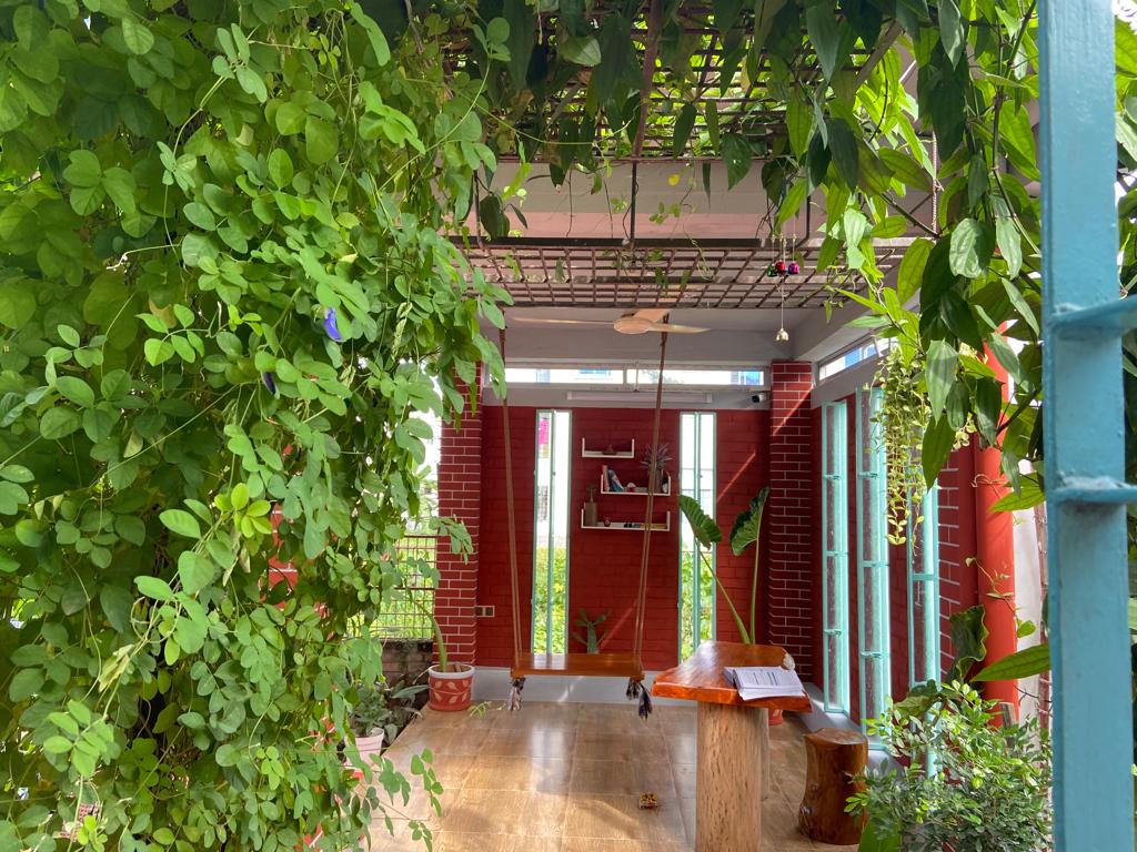 An outdoor space is framed by lush green vines, creating a natural canopy. The wooden floor extends under a structured roof, while bright red walls provide a vibrant backdrop. A small hanging lamp and minimalist decor enhance the inviting ambiance.