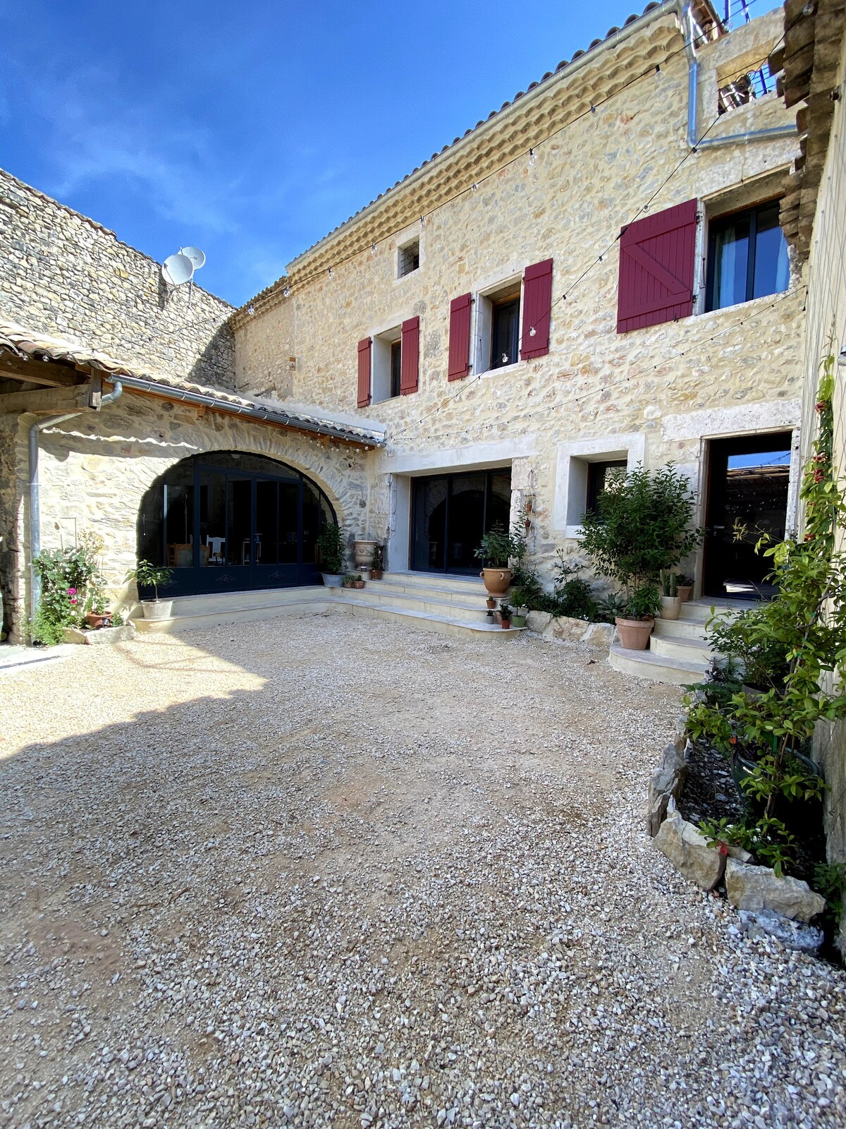 The historic stone building features a charming courtyard with gravel flooring and several potted plants. Large glass doors provide access to the interior, and red shutters frame the windows, creating a harmonious blend of rustic and modern elements under a clear blue sky.