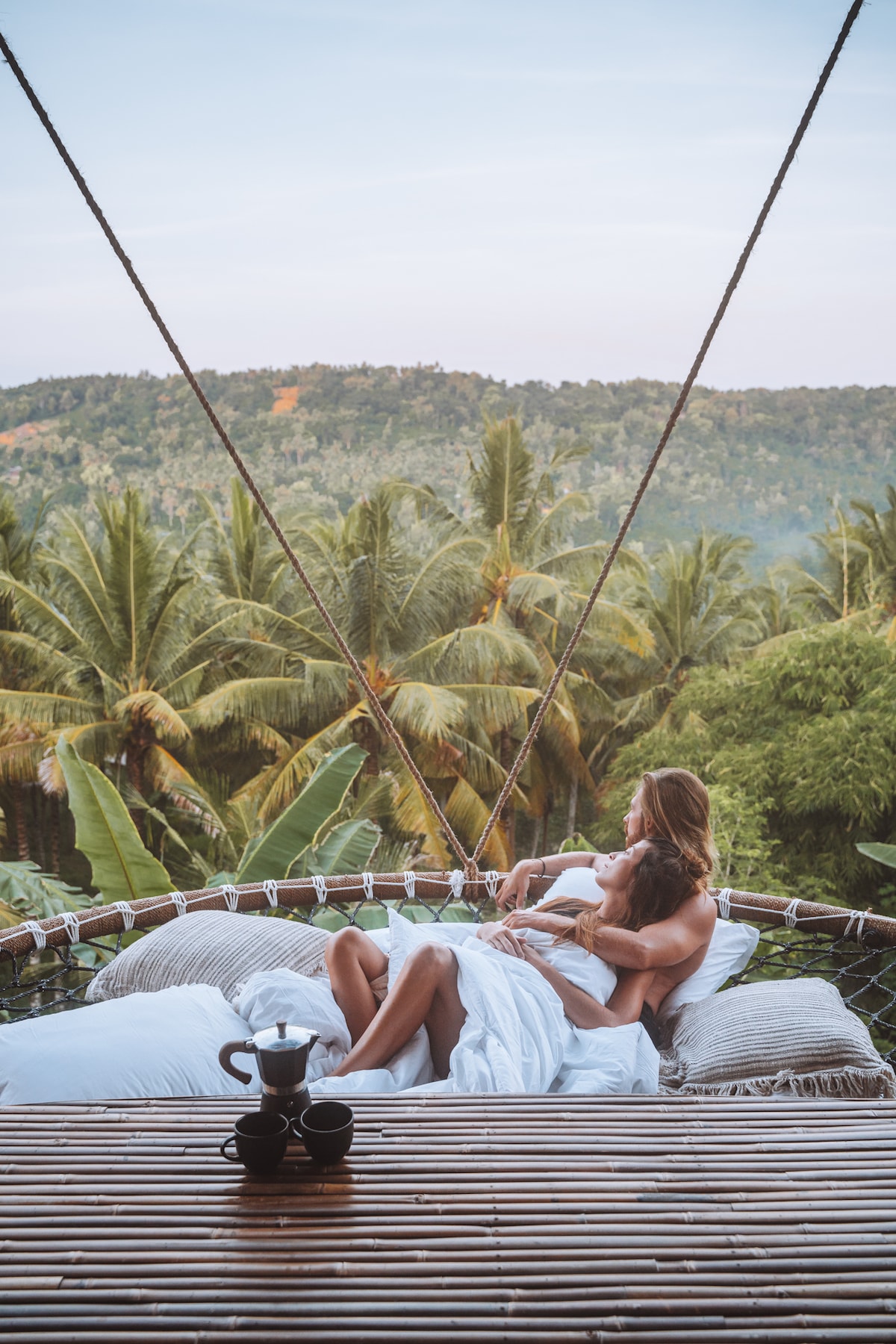A hammock is suspended between trees, overlooking a lush landscape filled with palm trees and greenery. Two people are seated on the hammock, surrounded by white bedding. A wooden table holds a coffee set, providing a tranquil setting amidst nature.