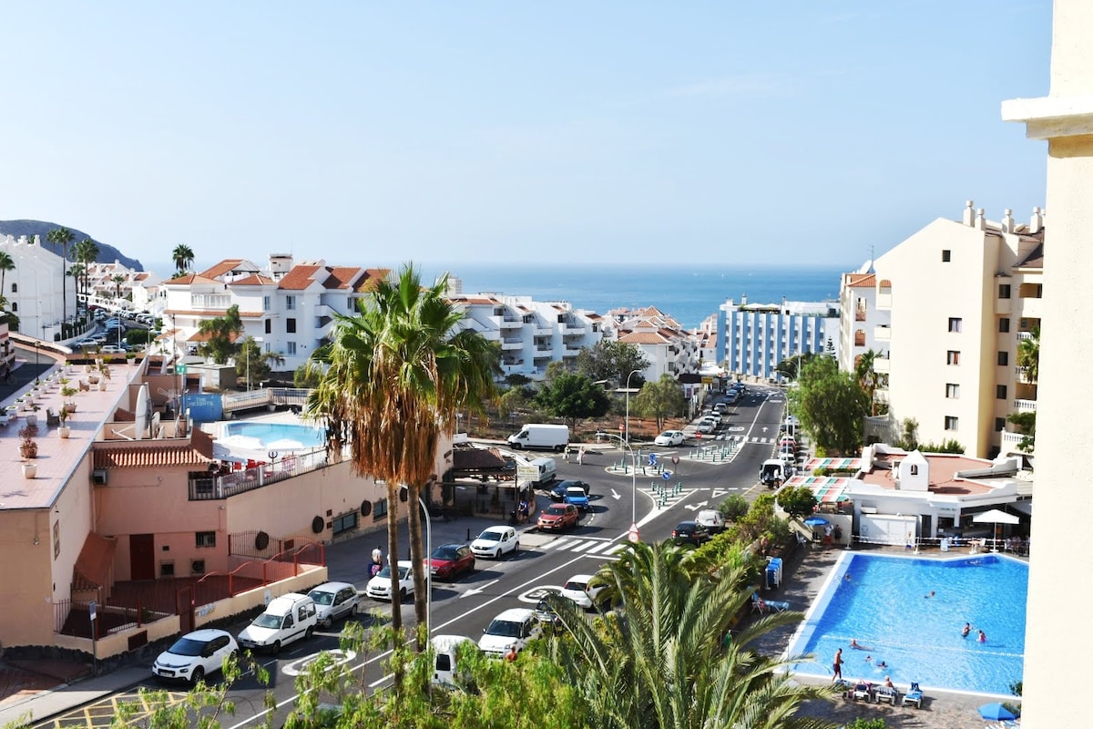 A scenic view captures the surrounding area, featuring buildings with a coastal backdrop. The image includes a glimpse of a turquoise pool and palm trees, enhancing the impression of a vibrant community. The ocean is visible in the distance, framed by the architecture.