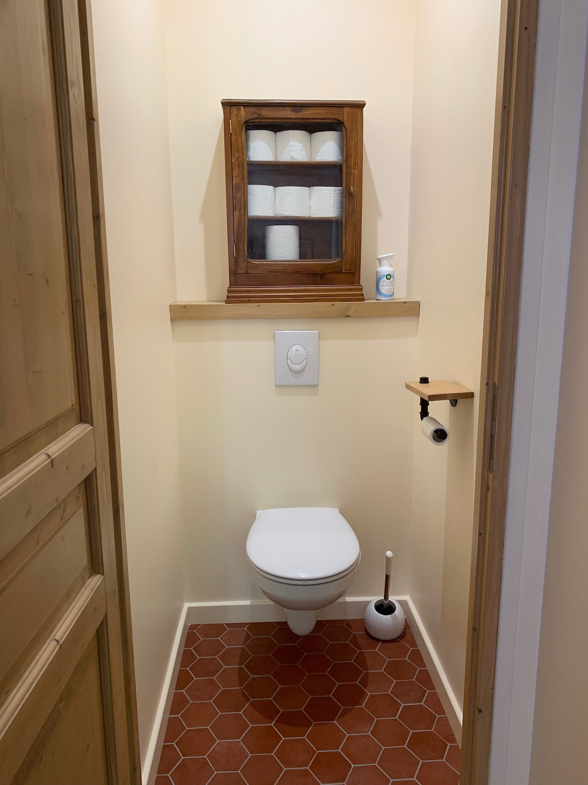 A compact bathroom is shown, featuring a clean white toilet positioned against a light-colored wall. Above, a wooden cabinet displays neatly stacked toiletries. A small shelf is attached next to the toilet, and a cleaning brush is placed nearby on the tiled floor.