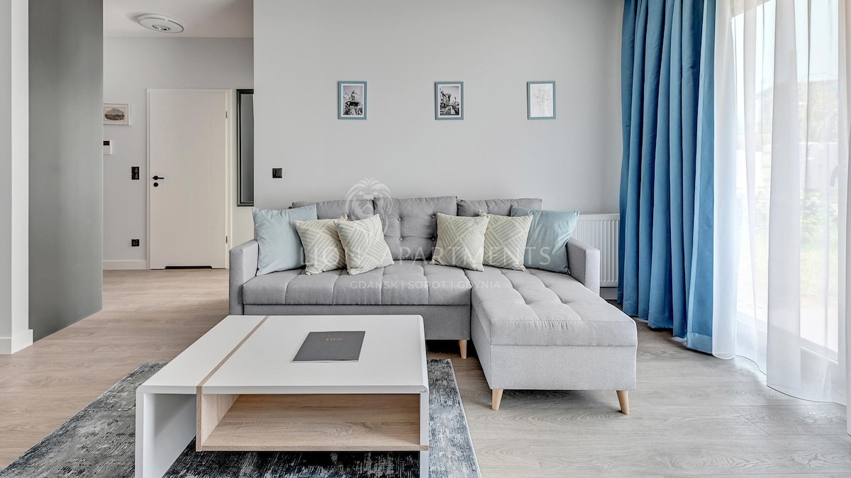 A bright living area features a comfortable gray sofa adorned with light-colored cushions. A modern coffee table sits in front, complemented by a soft area rug. Large windows dressed with light blue curtains allow natural light to fill the space.