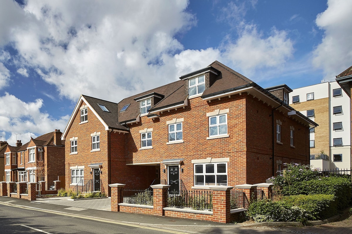 A row of modern brick buildings is seen along a quiet street, characterized by large windows and a welcoming entrance. The well-maintained landscape includes small hedges and a path leading to the front doors, with a backdrop of a bright, partly cloudy sky.