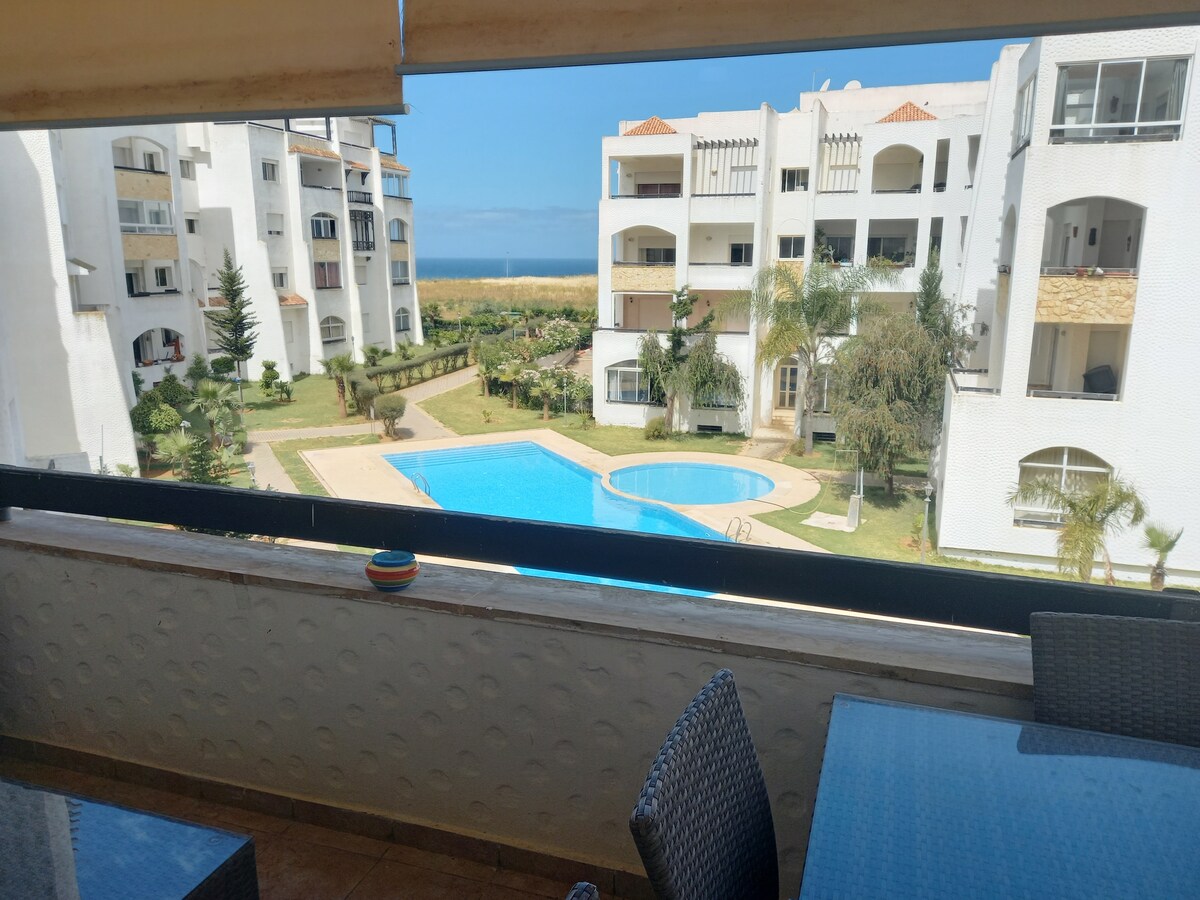 A scenic view is captured from the balcony, showcasing a clear blue pool surrounded by well-maintained gardens. In the background, the ocean is visible, enhancing the serene atmosphere of the residence. The balcony features a dining table set with wicker chairs.