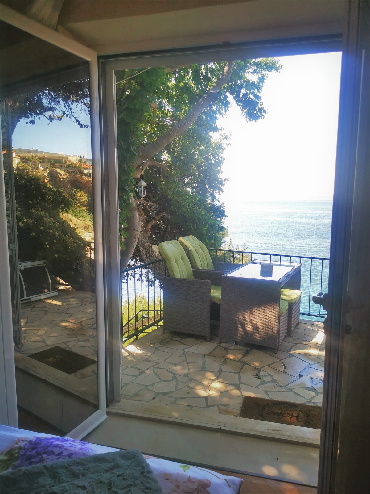 A bright terrace is visible through glass doors, featuring a seating arrangement with cushions overlooking the sea. The stone floor complements the lush greenery surrounding the area, creating a serene setting for relaxation.
