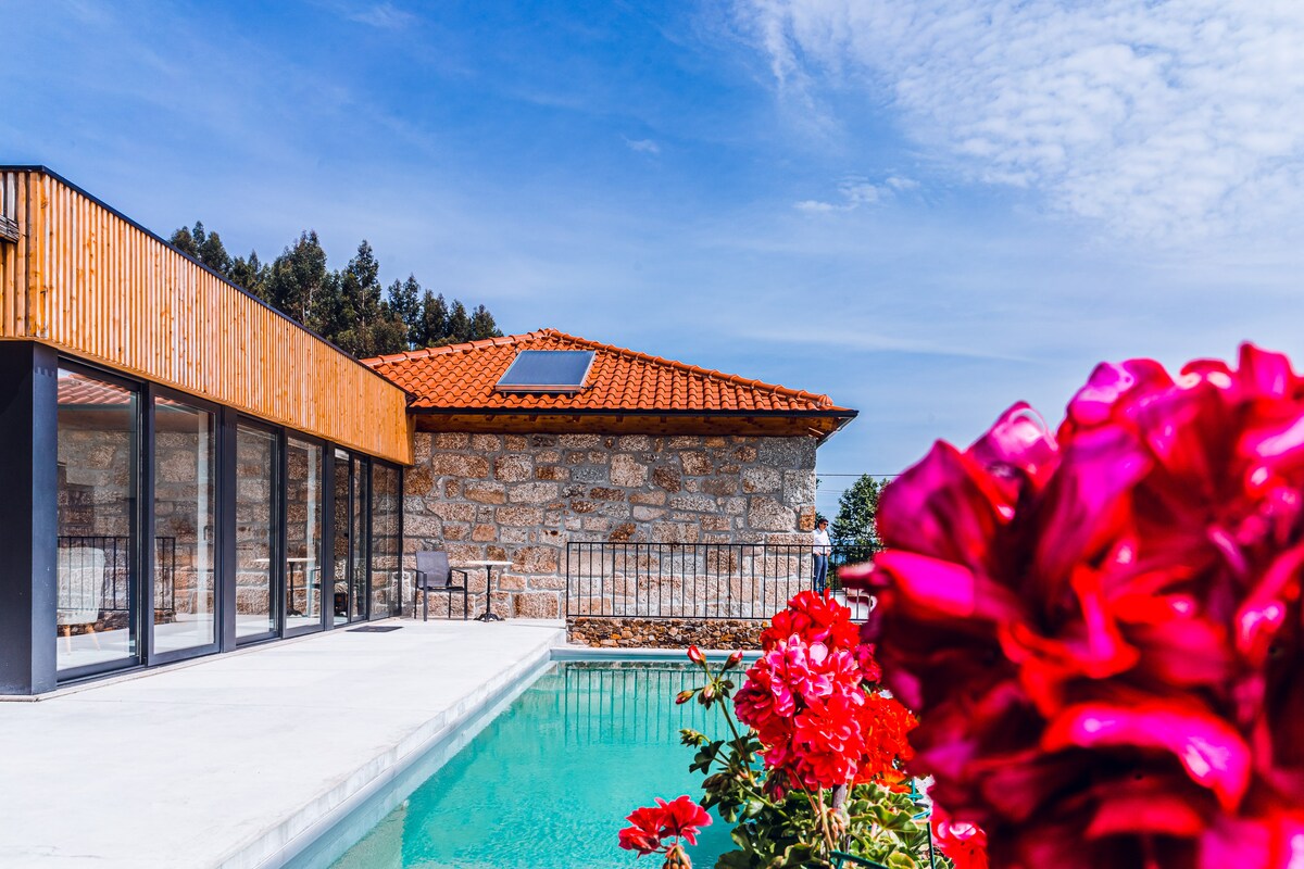 A modern structure with wooden accents is depicted beside a serene infinity pool. Colorful flowers frame the foreground, adding vibrancy to the scene. The clear sky enhances the overall bright atmosphere, while large glass doors open into the inviting outdoor space.