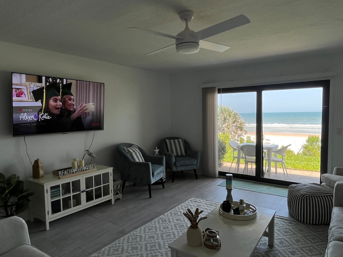 The living room features two armchairs and a white coffee table situated on a light area rug. A large television is mounted on the wall, while sliding glass doors provide a view of the beach and ocean beyond. Natural light fills the space.