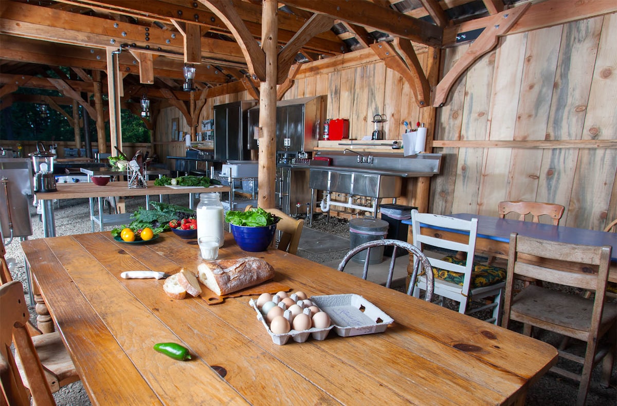 A spacious communal cookhouse features a large wooden table surrounded by assorted chairs. Fresh ingredients such as salads, vegetables, and eggs are displayed, with cooking equipment visible in the background. Natural light filters through the open design, creating an inviting cooking space.