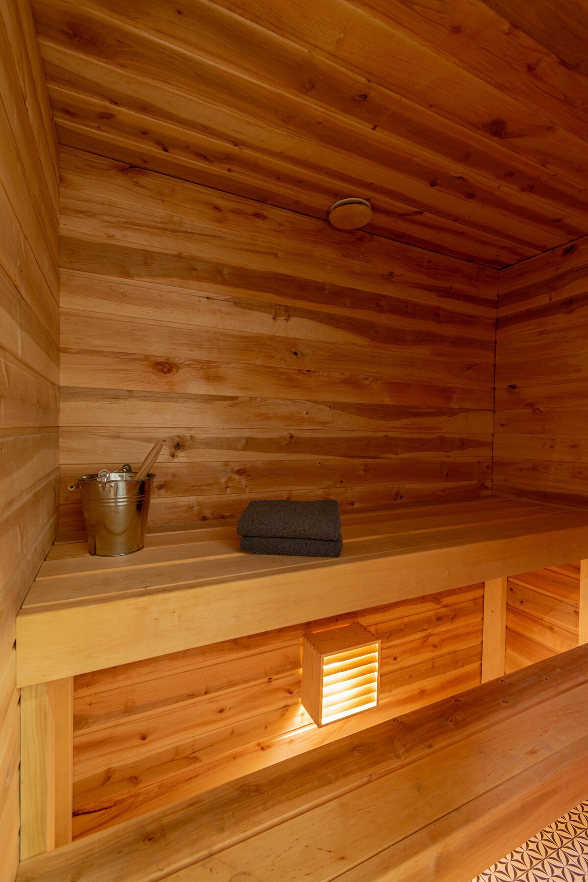The sauna features warm wooden walls and bench seating, providing a cozy environment for relaxation. A neatly folded gray towel is placed on the bench, alongside a small metal bucket. Soft lighting accents the space, creating a calming atmosphere.