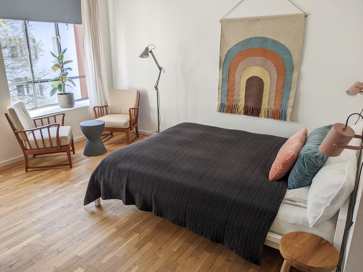 A bright and airy bedroom features a spacious bed with a dark blanket and decorative pillows. A wall tapestry adds a touch of color. Two armchairs, a small table, and a floor lamp are positioned near the window, allowing natural light to flow in.