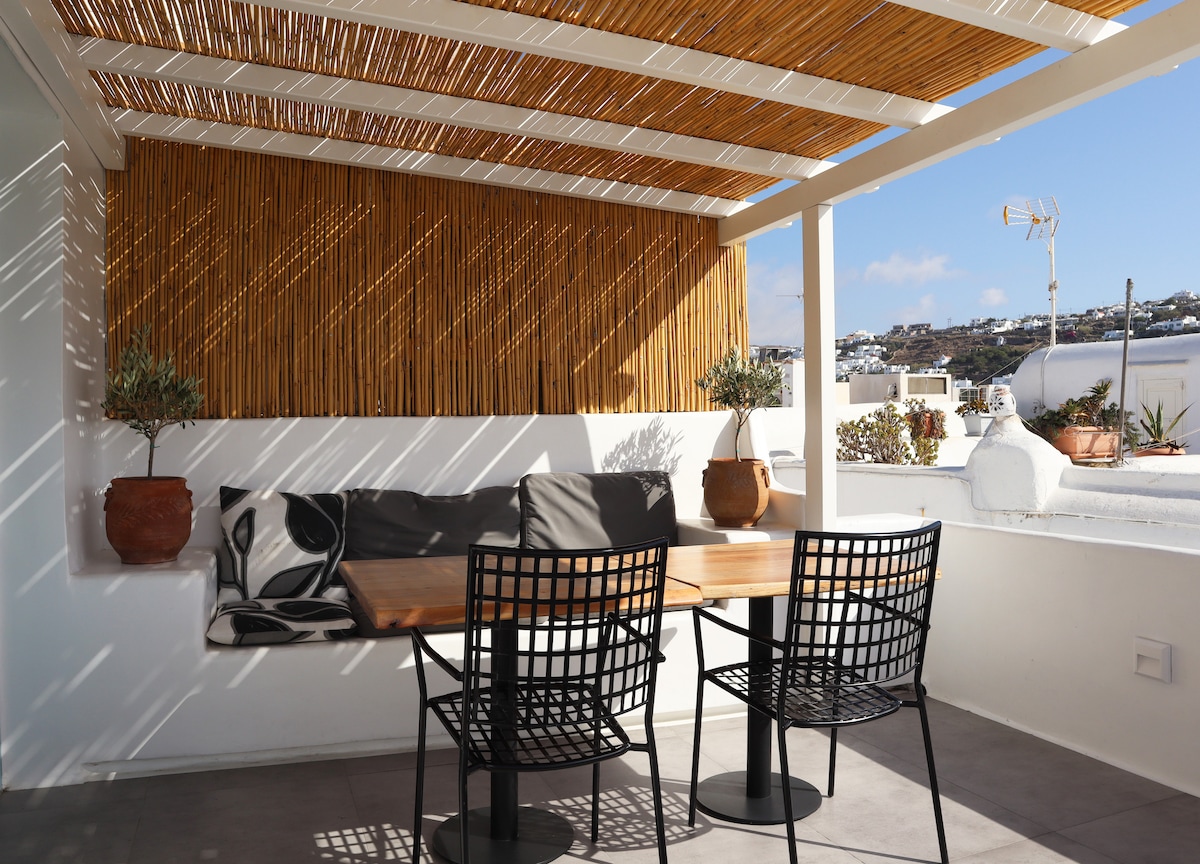 A private shaded rooftop terrace features a wooden overhang, creating a serene space. A wooden table is accompanied by two modern black wire chairs. Potted plants add greenery, while the surrounding walls offer privacy. Sunlight casts gentle patterns across the area.