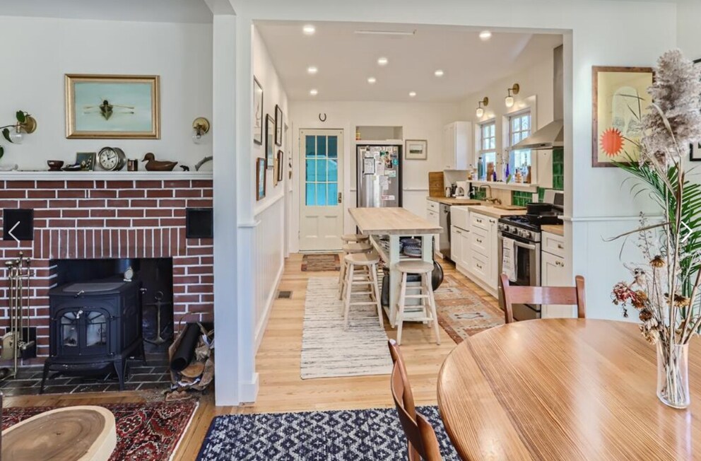 An open-floorplan kitchen and dining area features a long wooden table with ample seating. A brick fireplace is visible, adding an element of warmth. The kitchen showcases modern appliances and cabinetry, with natural light illuminating the space from multiple windows.