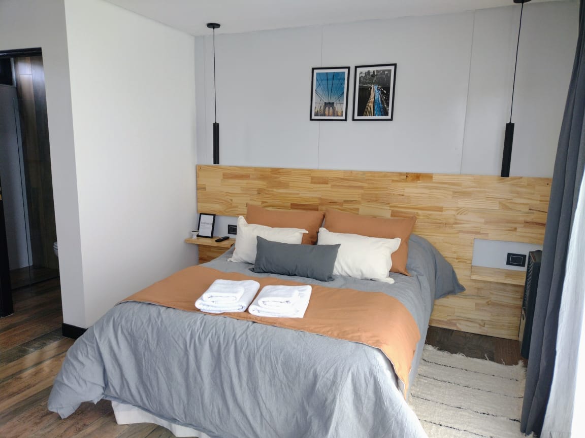 A cozy sleeping area features a spacious bed with neutral-colored linens and decorative pillows. The wooden headboard adds warmth, and framed photographs are displayed above. Soft light fixtures are suspended on either side, enhancing the modern design. A light-colored rug complements the wooden flooring.
