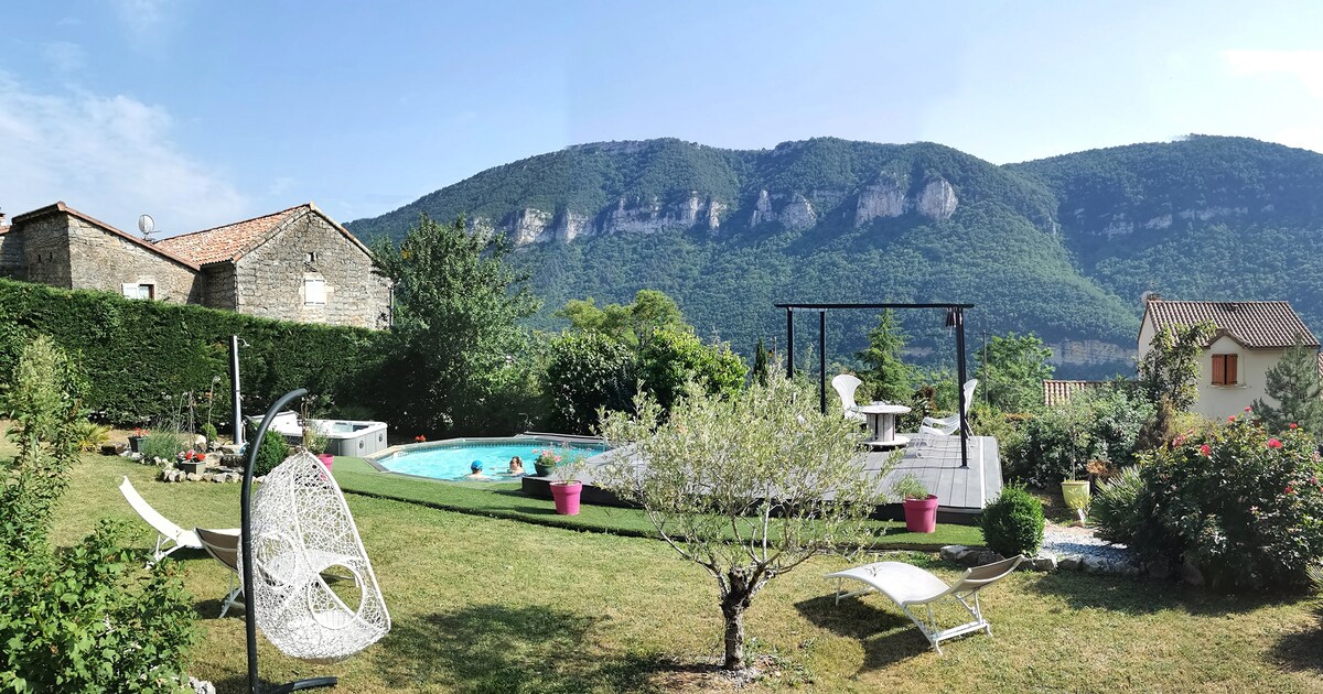 A serene outdoor area features a swimming pool surrounded by lush greenery and a wooden deck. Sun loungers are arranged around the pool, and vibrant flowers are visible along the landscape. Rolling hills rise in the background under a clear blue sky.