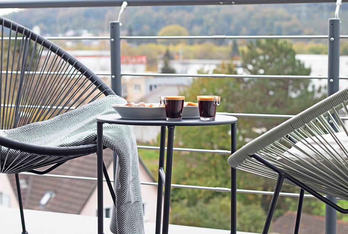 A balcony setting features two stylish chairs positioned around a small table. Two cups of espresso sit on a tray, accompanied by a selection of pastries. A cozy blanket drapes over one chair, with an expansive view of the greenery in the background.