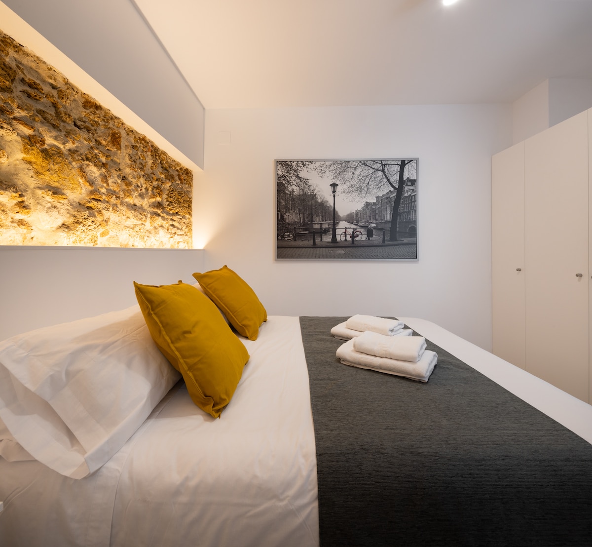 A modern bedroom features a spacious bed layered with light linens and decorative pillows. A textured stone accent wall provides a warm backdrop, while a black-and-white photograph of a city scene adds visual interest. Minimalist wardrobes are positioned to the side.