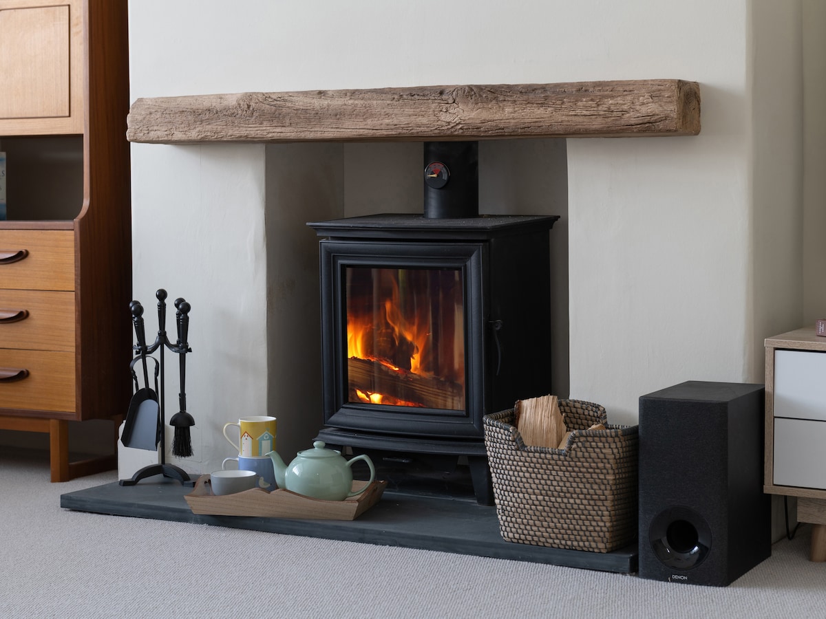 A wood-burning stove is featured prominently in the living area, set within a simple fireplace surround. A rustic wooden beam spans above, and a selection of firewood sits nearby. Additional items include a green teapot, mugs, and a woven basket, creating a cozy setting.