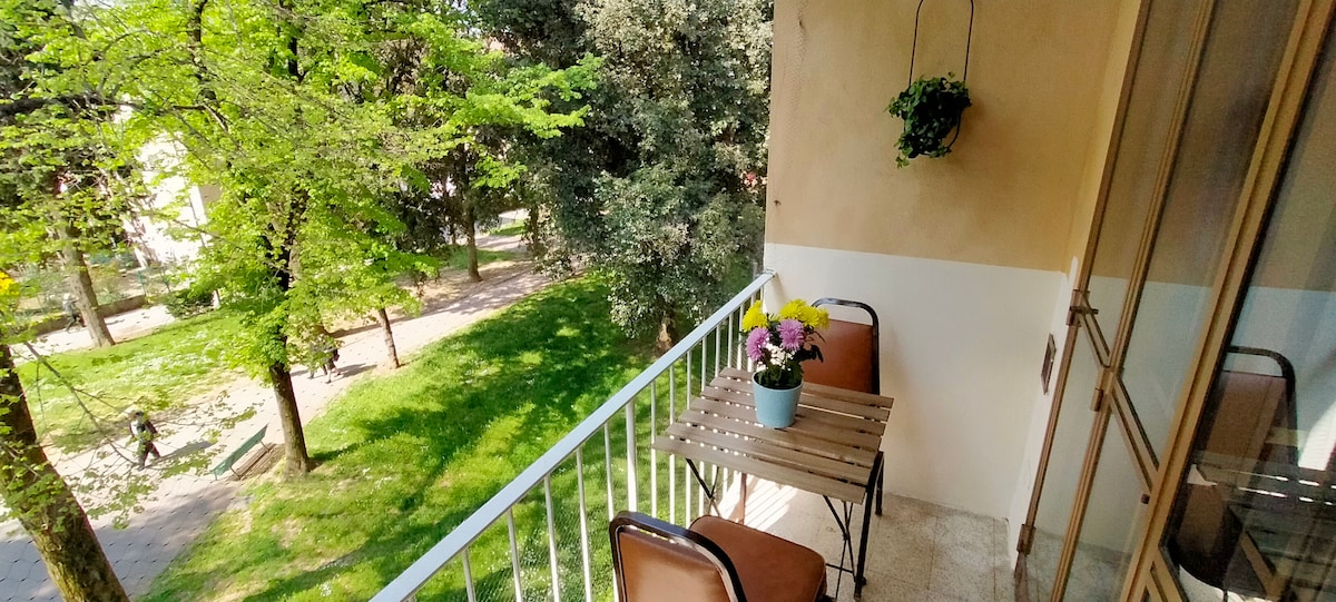 A small balcony is shown, featuring a wooden table with two chairs. A potted plant holds colorful flowers atop the table. Lush greenery is visible in the background, with walking paths and trees enhancing the outdoor ambiance.