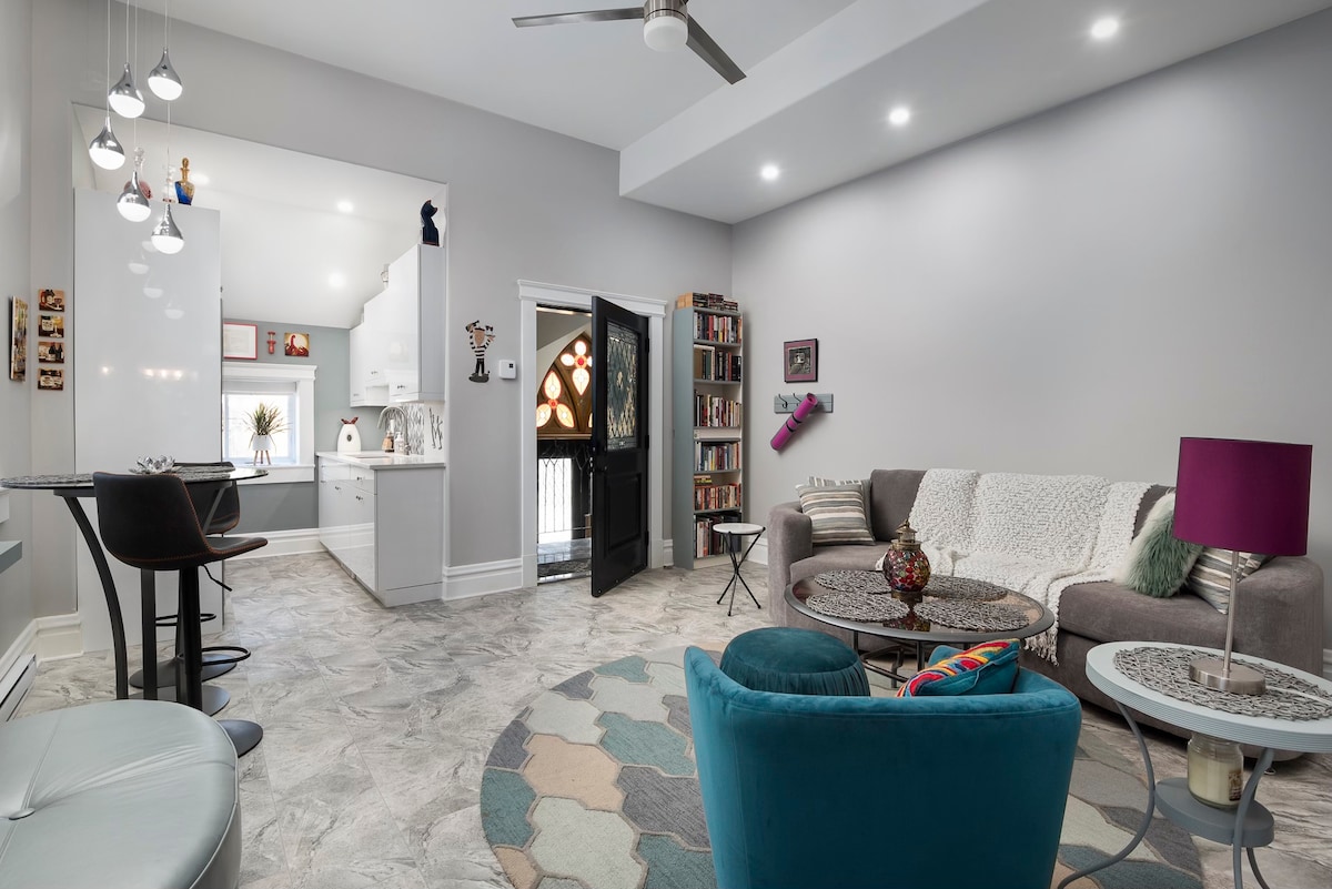 A modern living area is visible, featuring a comfortable sofa and colorful accent chairs arranged around a stylish coffee table. Natural light enters through a nearby doorway leading to the kitchen. Decorative light fixtures hang from the ceiling, and a bookshelf adds character to the space.