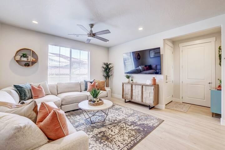 A spacious living area features a large, comfortable sectional sofa adorned with decorative pillows, complemented by a round coffee table. A 75" smart TV is mounted on the wall, opposite a window with natural light filtering through. Decorative shelves and a console table add style and storage.