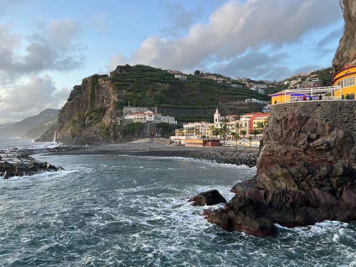 The image captures a rugged shoreline with the Atlantic Ocean's waves gently lapping against the rocks. In the background, lush green hillside terraces are visible along with colorful buildings of the village, creating a harmonious blend of nature and architecture.