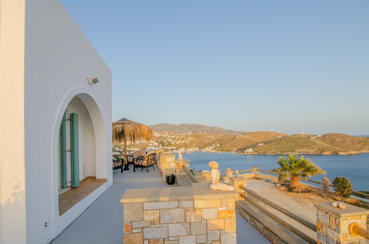 An outdoor terrace is presented, featuring a thatched umbrella and a seating area with dark wicker chairs. Scenic hills and the sea are visible in the background, while the structure showcases a white façade and a large arched window, offering ample natural light.