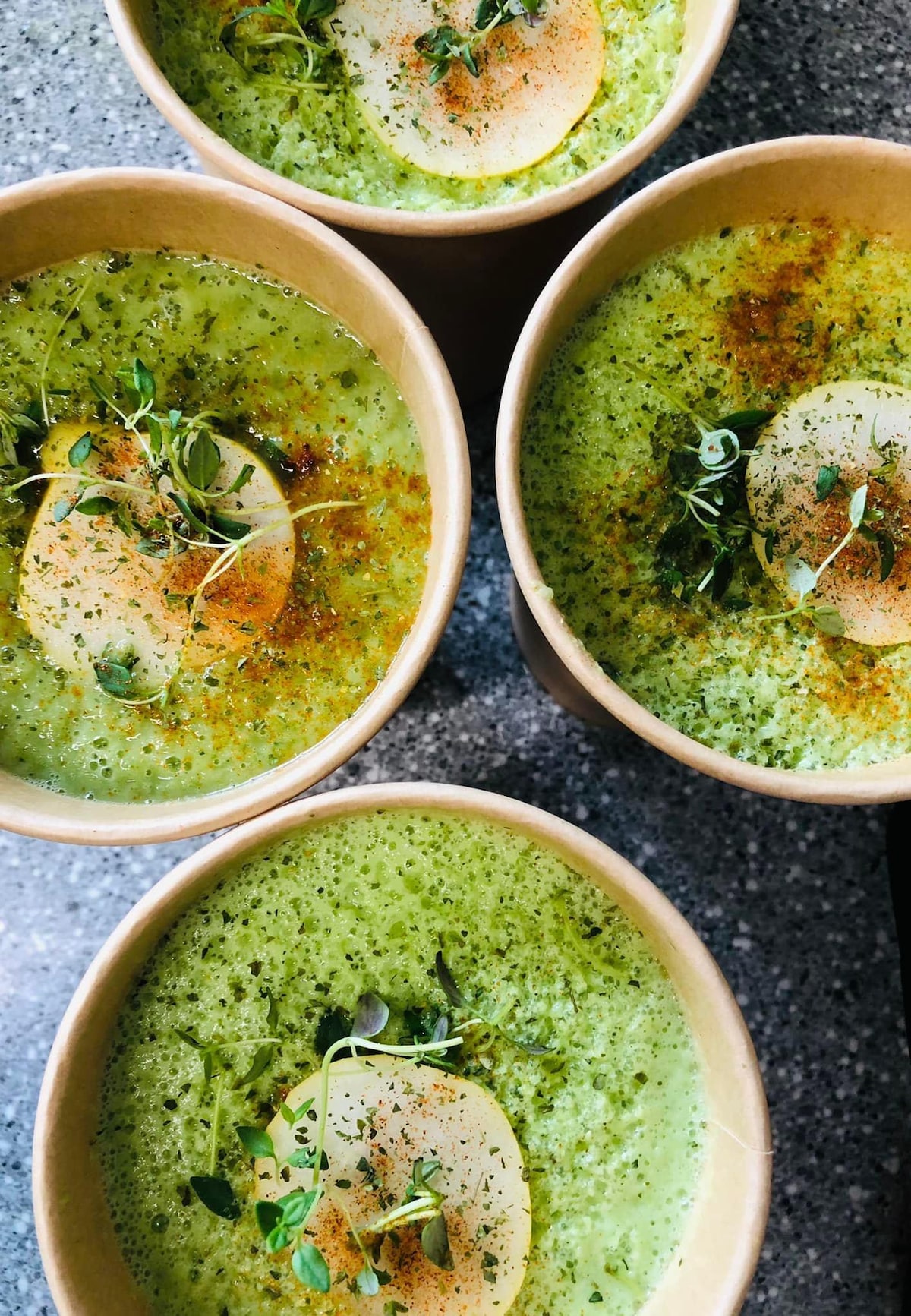 Four bowls of vibrant green soup are arranged closely on a countertop. Each bowl is garnished with a slice of pear, fresh herbs, and a sprinkle of spices, showcasing a healthy and nourishing meal option.