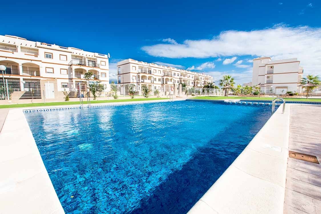A large communal pool is surrounded by well-maintained green spaces, providing a serene environment for relaxation. The water's clear blue surface reflects the sky, enhancing the inviting atmosphere. Modern apartment buildings are visible in the background, showcasing a blend of comfort and leisure.