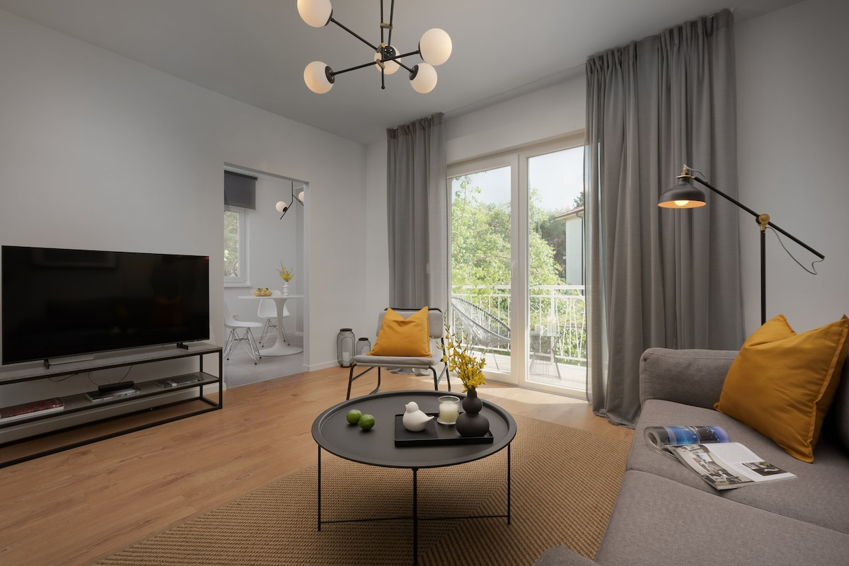 A modern living room is highlighted by a neutral palette, featuring a gray sofa adorned with yellow pillows. A stylish coffee table with decorative items sits at the center. Large windows invite natural light and offer views of the greenery outside.