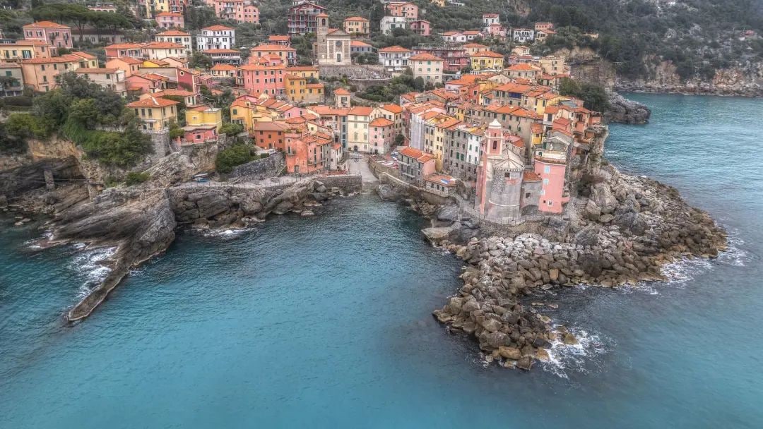 A coastal village is set against a backdrop of steep cliffs and azure waters. Colorfully painted buildings line the shoreline, creating a charming visual contrast with the rocky landscape. The beach area is visible, blending sand and stones where the sea meets the land.