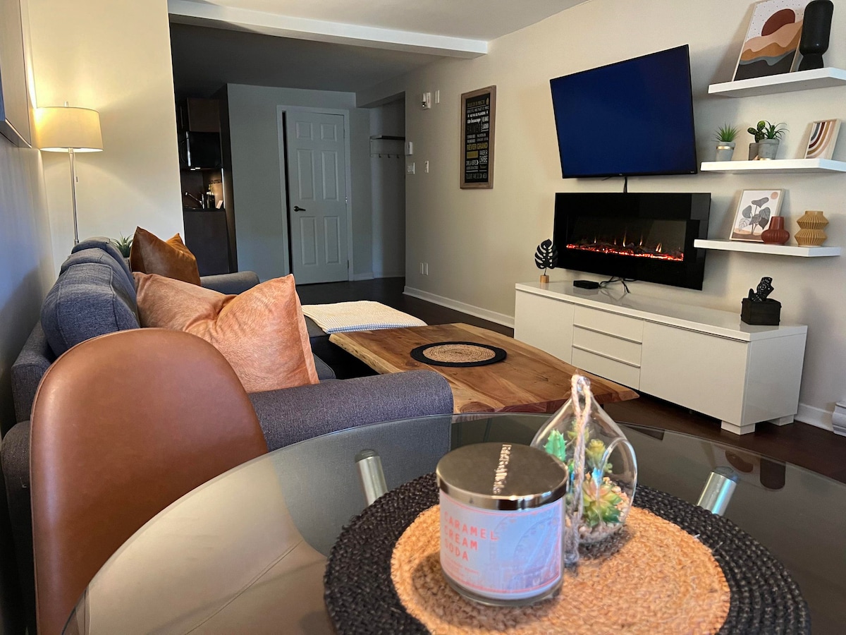 A spacious living area features a comfortable sectional sofa adorned with decorative cushions. A modern electric fireplace is mounted on the wall beneath a large television. A dining table is set nearby, showcasing a candle and decorative plants, contributing to the overall welcoming ambiance.