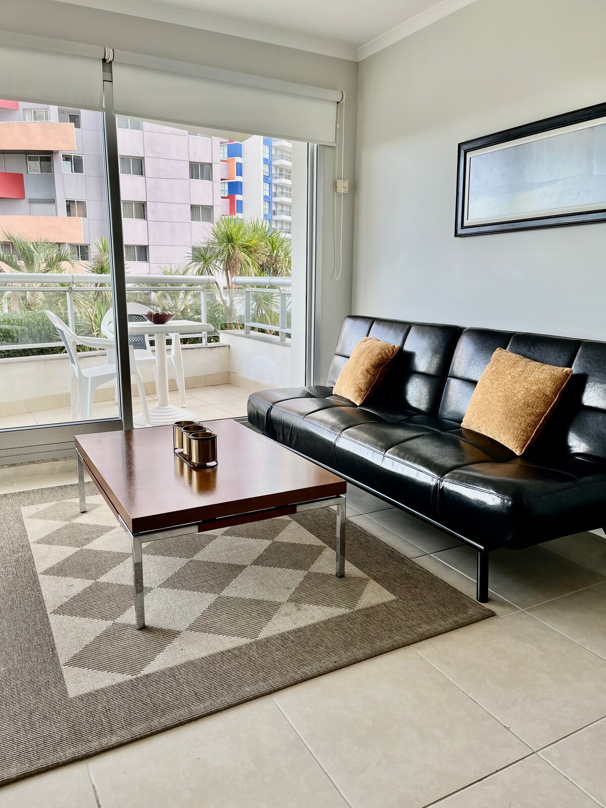 A modern living area features a sleek black sofa adorned with two decorative cushions. A wooden coffee table is positioned in front of the sofa, resting on a geometric patterned rug. Large glass doors lead to a balcony with outdoor seating.