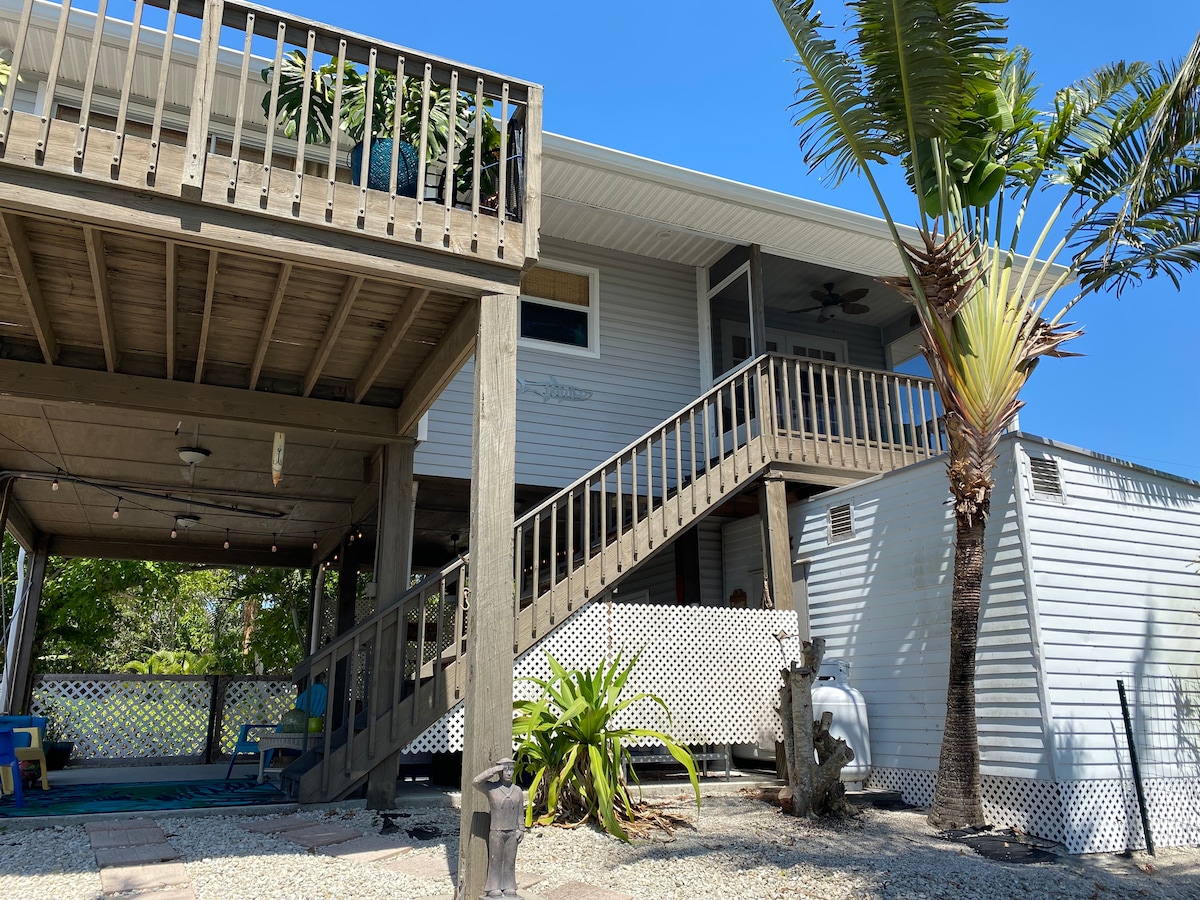 A two-story retreat is showcased, featuring a wooden deck with a staircase leading to the upper level. Lush palm trees and potted plants are visible, with a gravel landscape adding texture to the outdoor space.