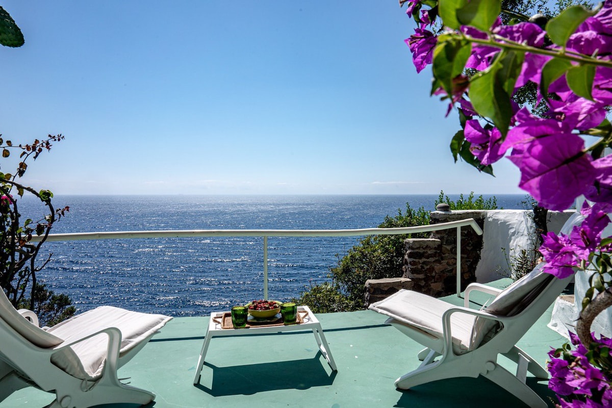 A serene terrace provides stunning views of the sea, framed by vibrant pink flowers. Two lounge chairs are positioned around a small table, ideal for relaxation. The expansive ocean horizon stretches beyond, reflecting a clear blue sky.