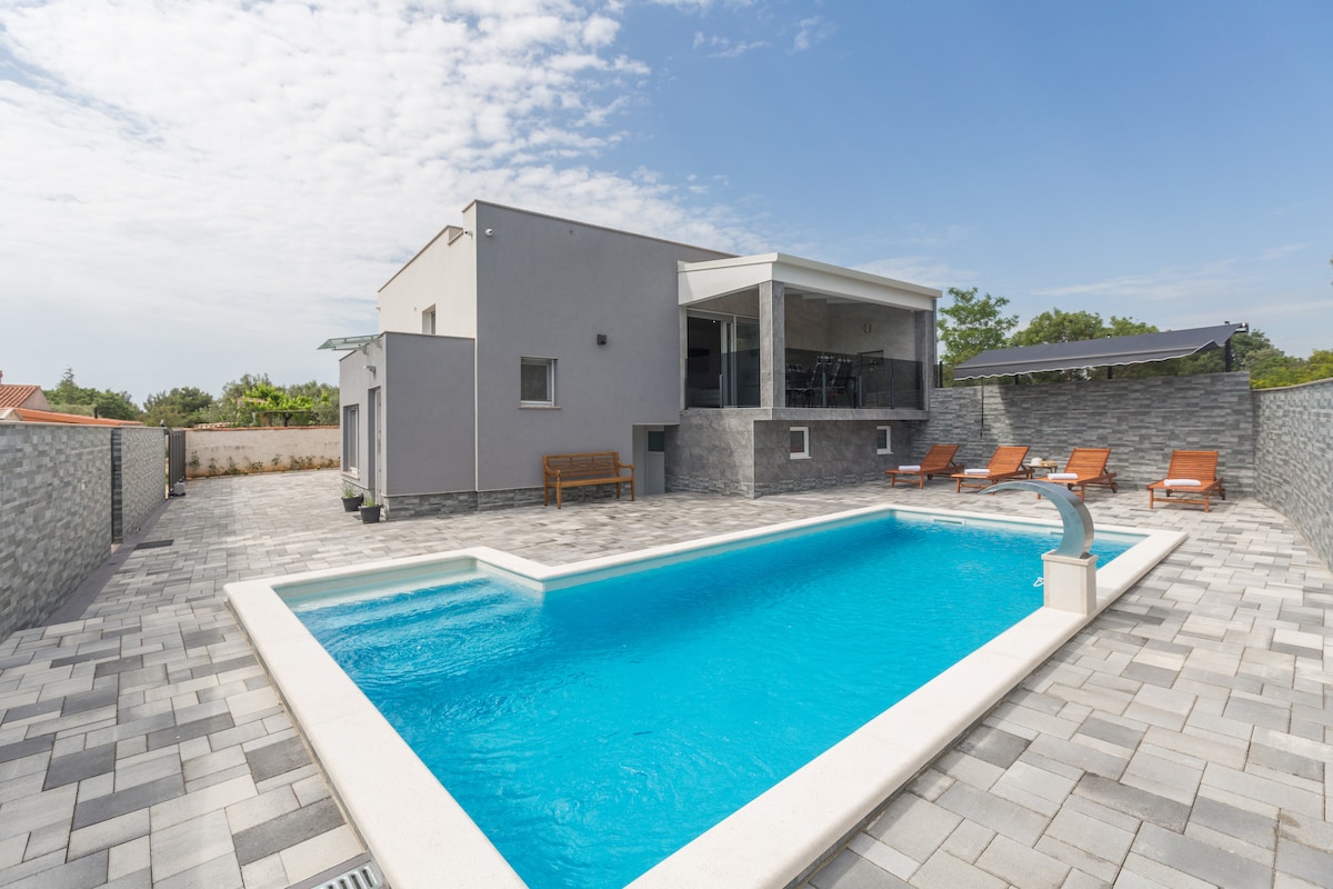 A modern villa is pictured with a clean-lined swimming pool at the forefront. Sun loungers are arranged beside the pool, and the exterior walls are clad in gray stone. A covered terrace can be seen on the upper level, providing a shaded area for relaxation.
