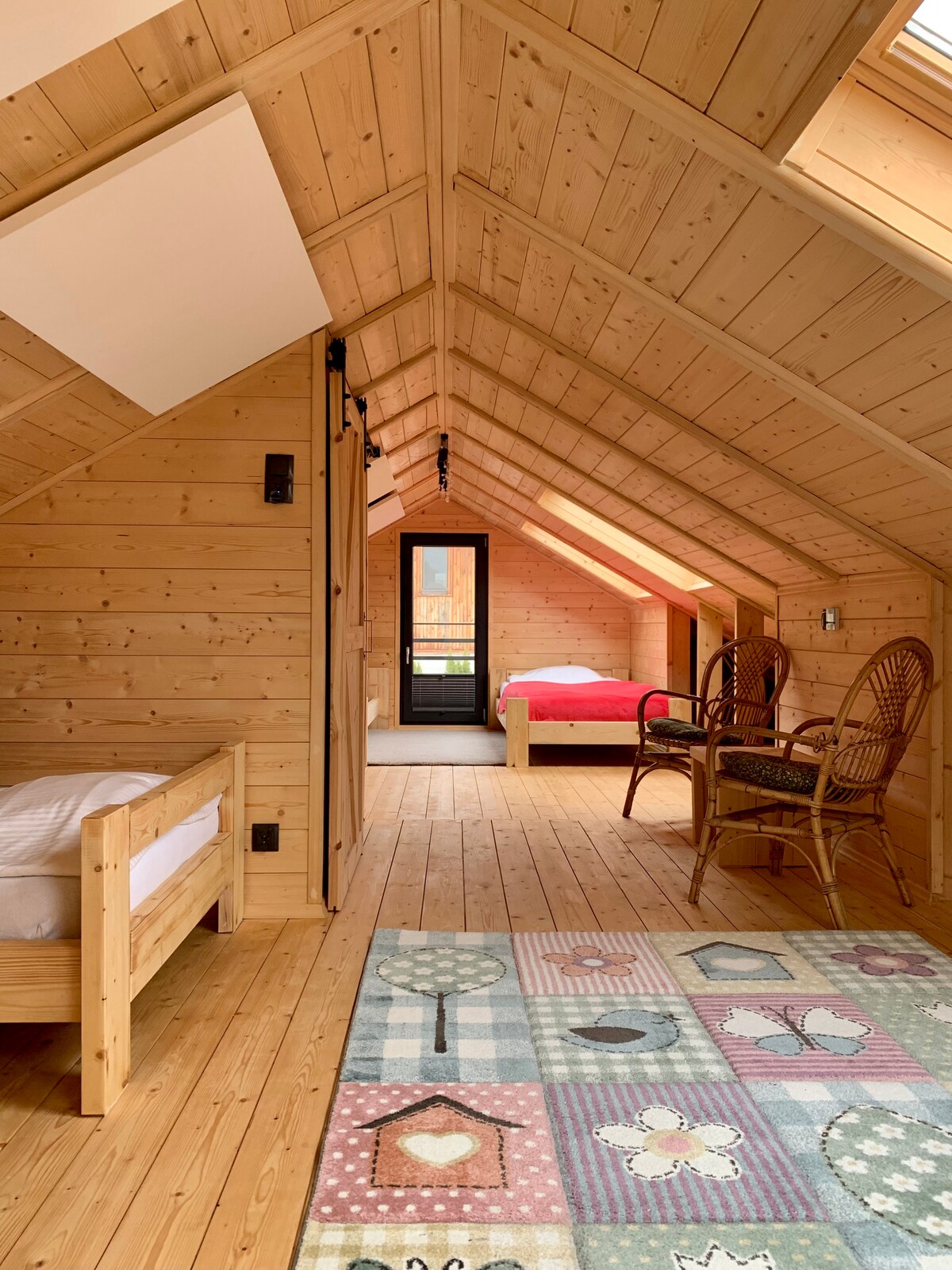 An inviting interior features a triangular wooden ceiling with natural light filtering through skylights. Two beds, one dressed in red, are situated near a door leading to a balcony. A patterned rug adds color to the wooden floor, complemented by two wicker chairs in the corner.