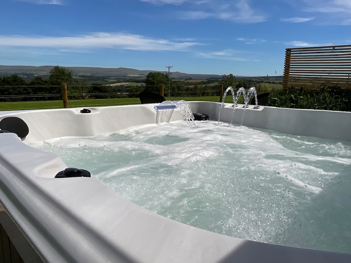 A spacious hot tub filled with bubbling water is positioned against a backdrop of open countryside. Water features are visible, with jets creating a relaxing ambiance. The clear blue sky and rolling hills enhance the tranquil outdoor setting.