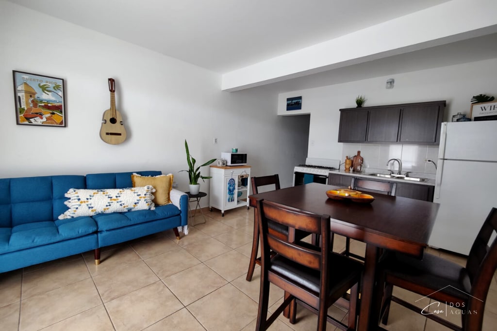 An open living space features a blue sofa adorned with cushions, a wooden dining table, and four chairs. A kitchenette with modern appliances is visible in the background. A wall-mounted guitar decorates the light-colored wall, complemented by a framed artwork. Natural light brightens the area.