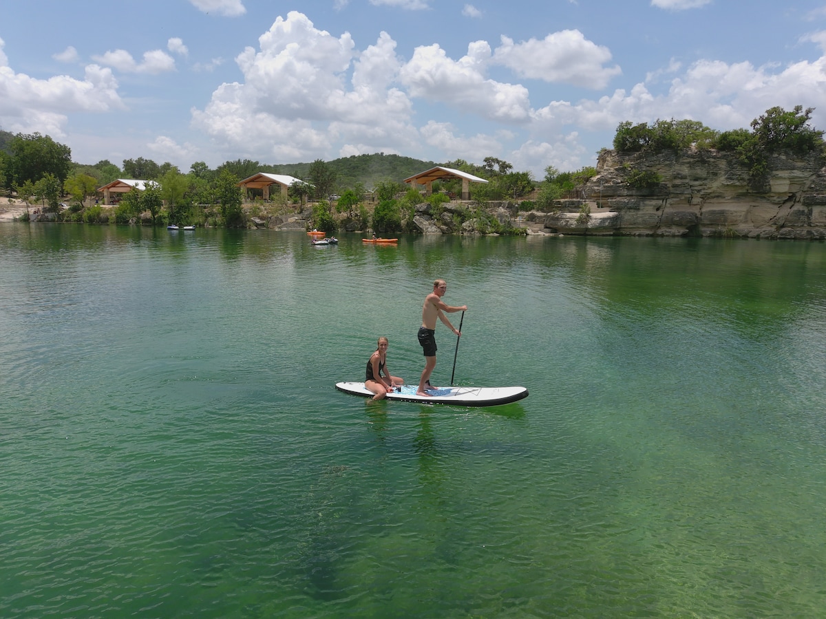 A paddle board is shown gliding over clear green waters, with two individuals enjoying the activity under a bright sky. In the background, several cottages are visible among lush greenery, complementing the serene river landscape.