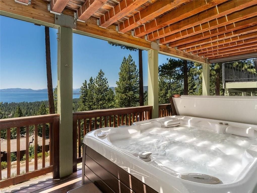 The hot tub, equipped with multiple jets, is positioned on a deck with panoramic views of Lake Tahoe. Tall trees surround the area, providing privacy and a natural setting. The clear blue sky complements the serene atmosphere and enhances the relaxing experience.