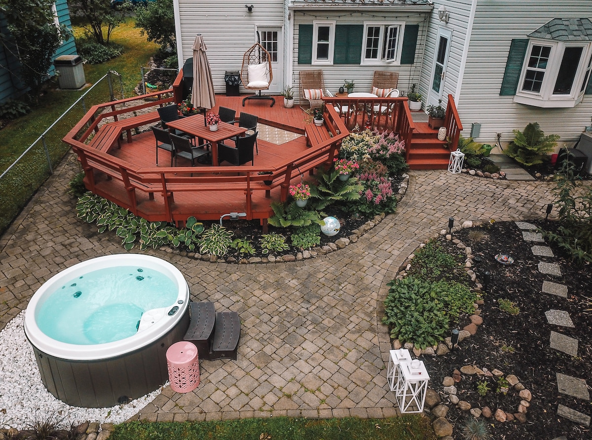 An outdoor space features a wooden deck adorned with seating and a dining table, shaded by an umbrella. A hot tub is positioned near landscaped gardens and a stone path, surrounded by vibrant flowers and greenery, creating a serene backyard ambiance.