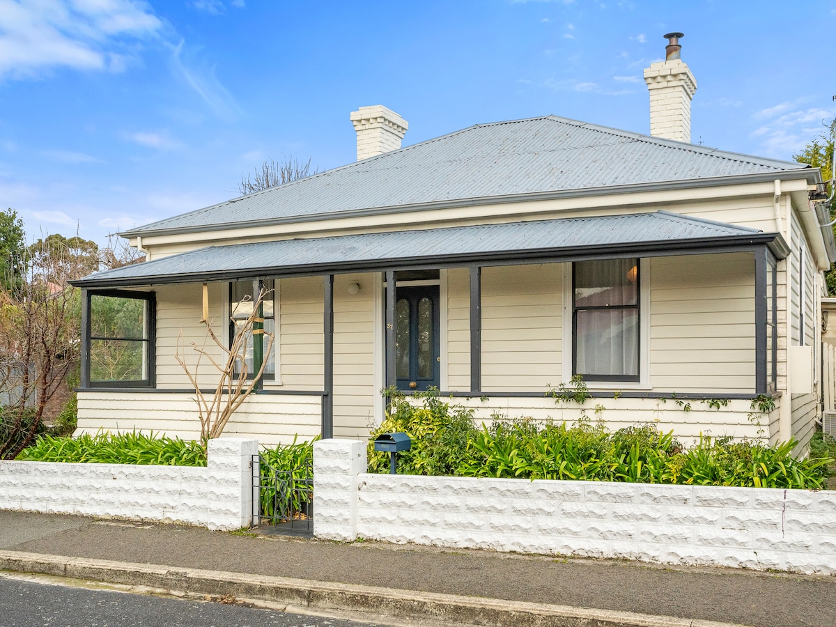 The charming cottage is framed by lush greenery and a white stone fence, highlighting its inviting facade. A covered porch with a welcoming entrance is present, and the roof features a classic design typical of the area. Soft colors complement the serene environment.