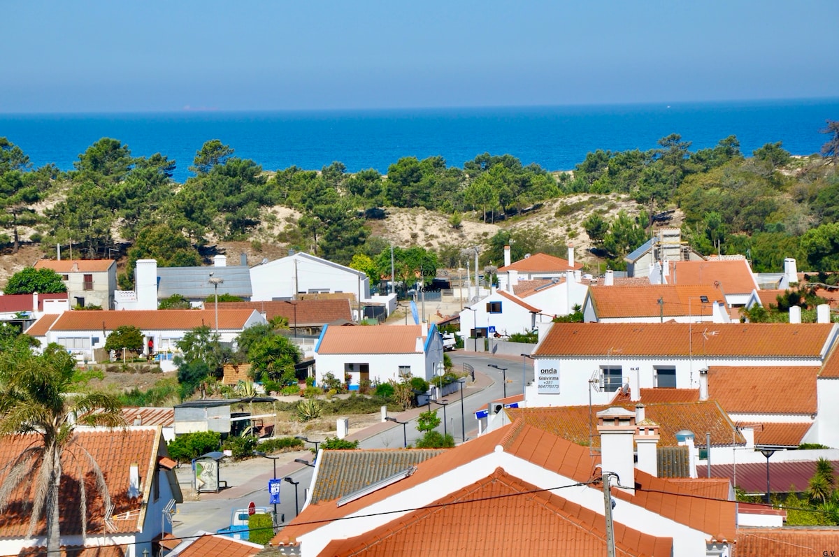 A panoramic view showcases a coastal town with white-roofed buildings and terracotta roofs. Lush greenery and sandy areas are visible in the background, leading to a tranquil blue sea under a clear sky.